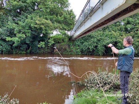 Hunsingore. Martin does a spectacular long-distance sample from the bank of the river Nidd above a footbridge. The river had been over the bank recently, after heavy rain. Chocolate coloured river with a line of foam in the centre of the river.